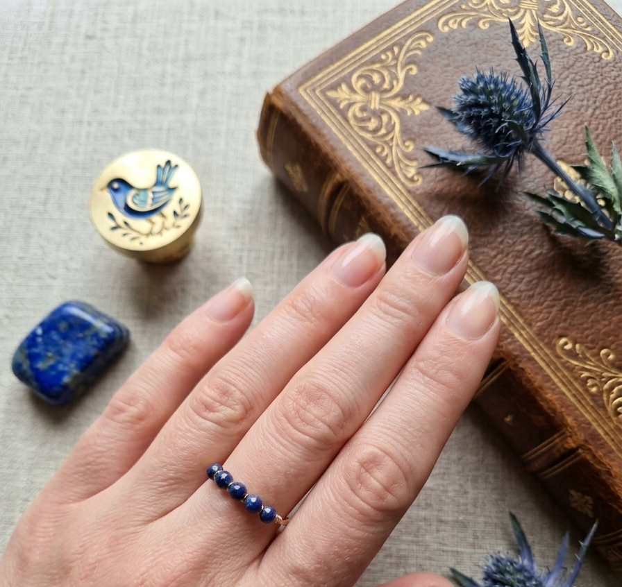 Käsi, jossa on tummansinisistä helmistä tehty sormus, lepää kirjan vieressä. Kuvassa näkyy myös sininen thistle‑kukka, sininen kivi ja kultainen riipus, jotka luovat harmonisen boho‑henkisen asetelman.
